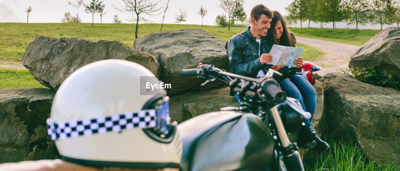 Panoramic shot of smiling bikers sitting on rocks while holding map on land