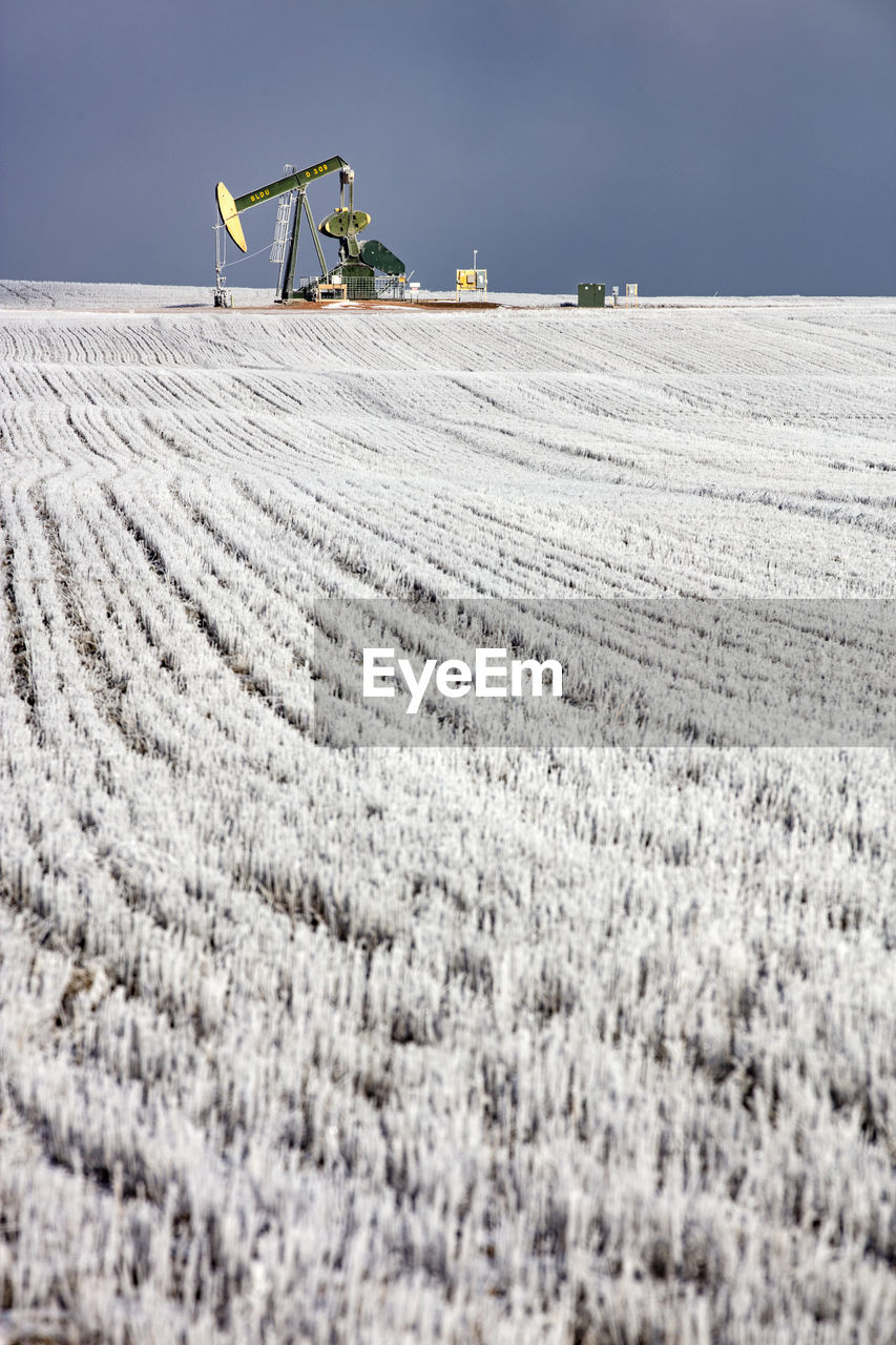 Mid distance view of pumpjack at oil industry on field against sky during winter