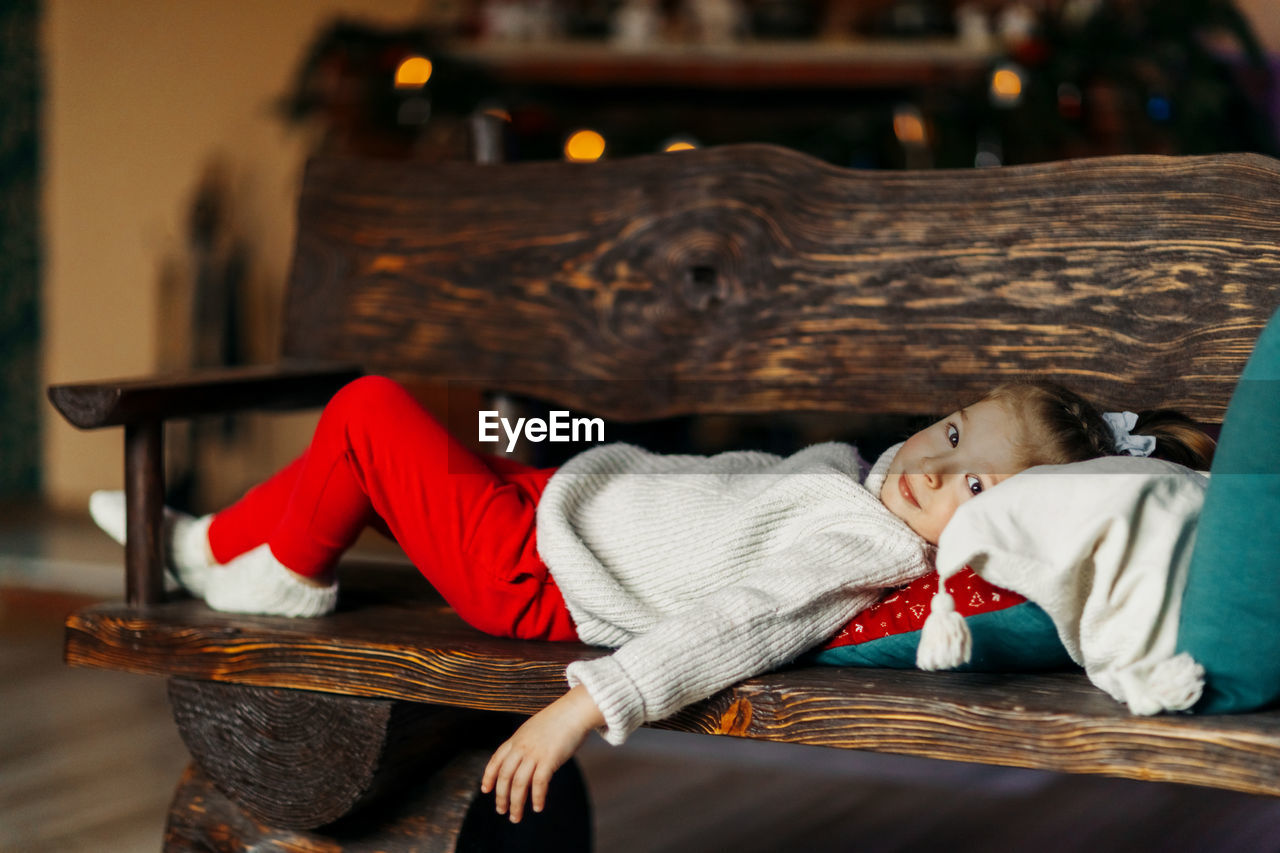 Little girl lies on a bench against the background of the fireplace and looks into the camera