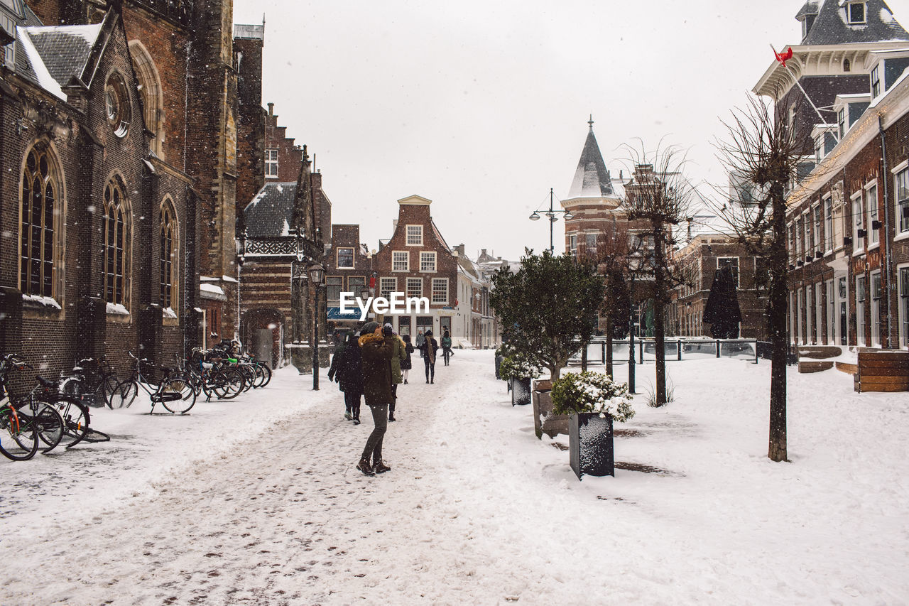 REAR VIEW OF PEOPLE WALKING ON SNOW COVERED STREET IN CITY