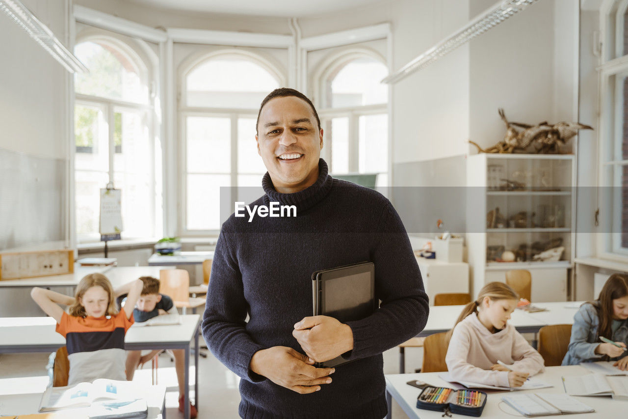 Portrait of smiling male teacher holding tablet pc while standing in classroom