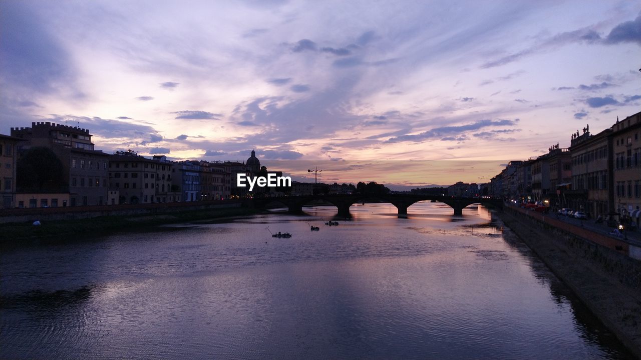 Bridge over river by buildings against sky at sunset