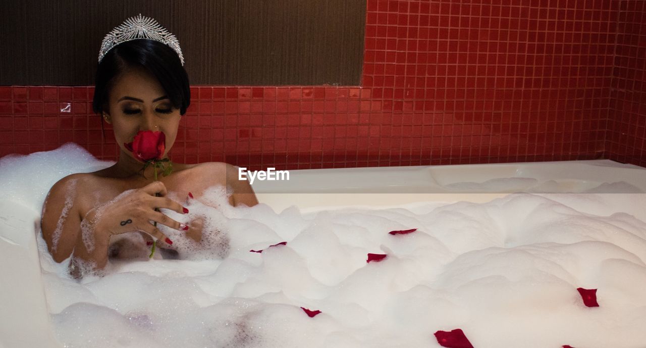 Shirtless woman smelling red rose while taking bubble bath in bathtub