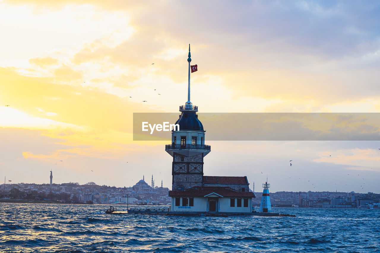 LIGHTHOUSE BY SEA AGAINST BUILDINGS DURING SUNSET