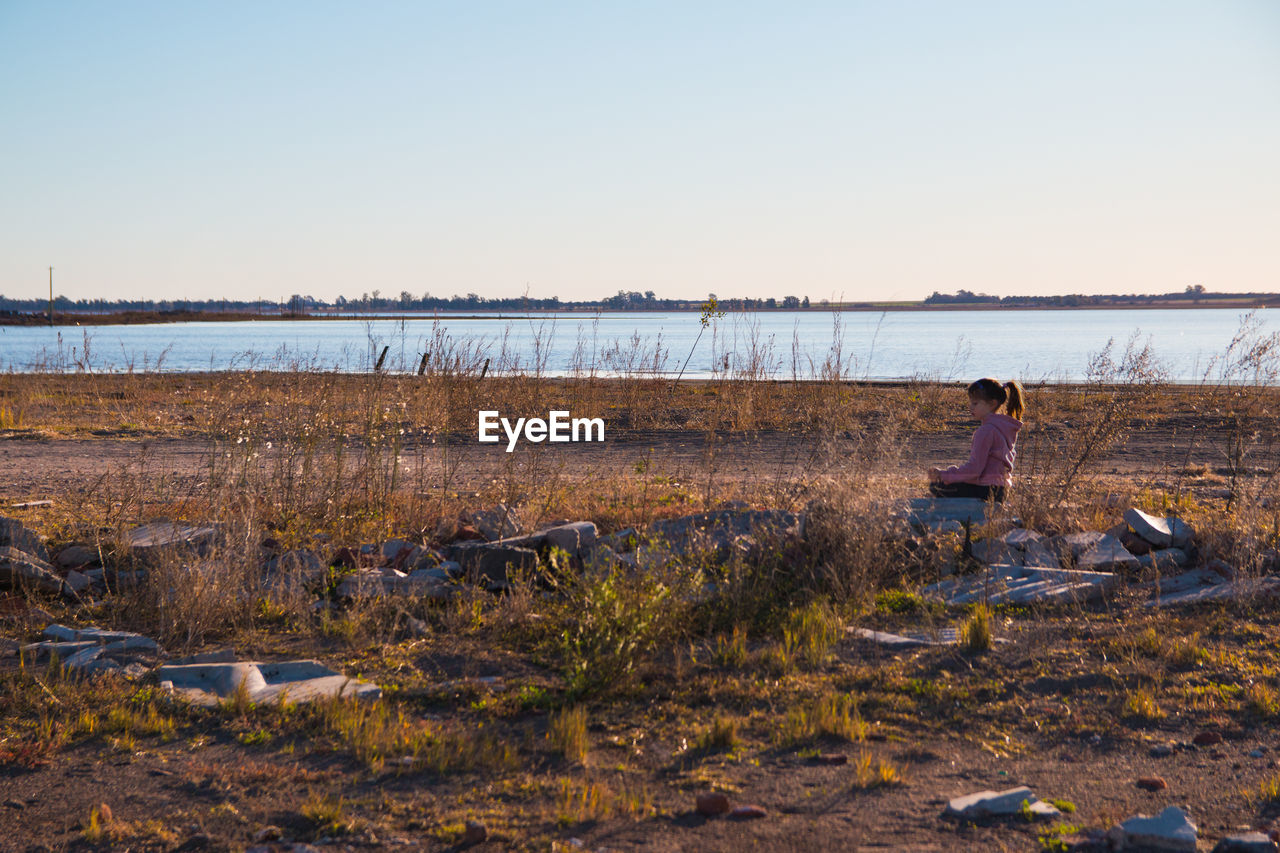 MAN SITTING ON SHORE AGAINST CLEAR SKY