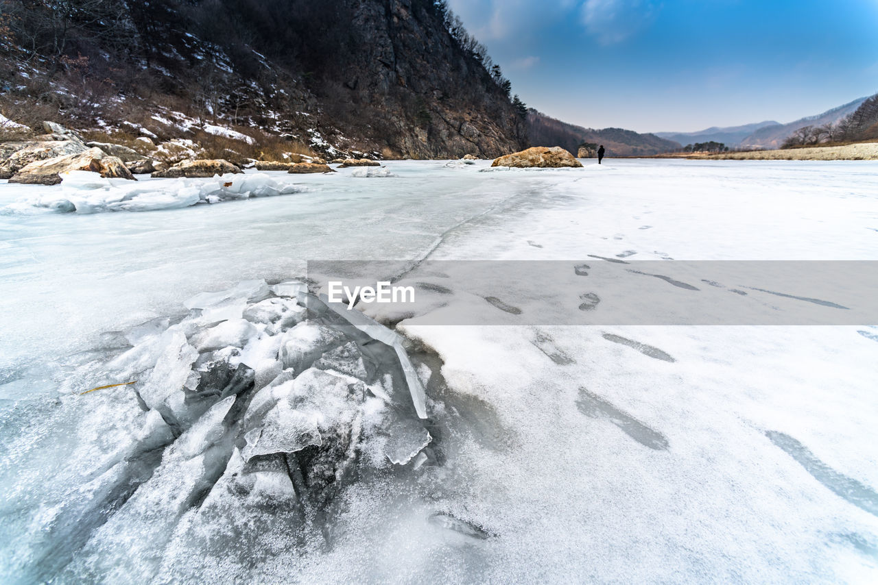 Frozen river by cliff against cloudy sky