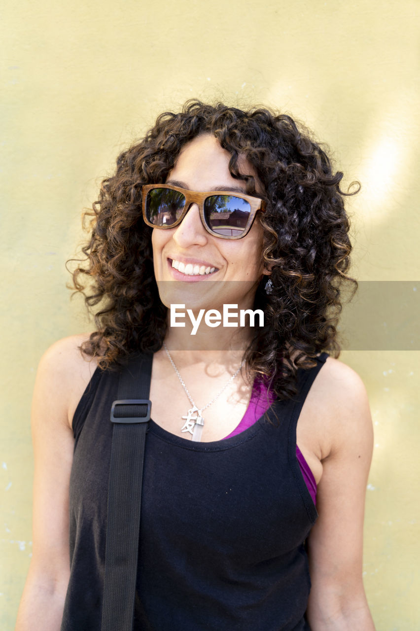 Mixed race woman with sunglasses standing against a yellow wall.