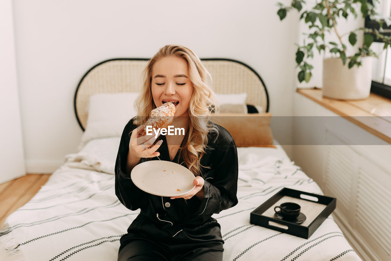 A happy pretty young woman in pajamas is resting and having breakfast in bed in a cozy apartment