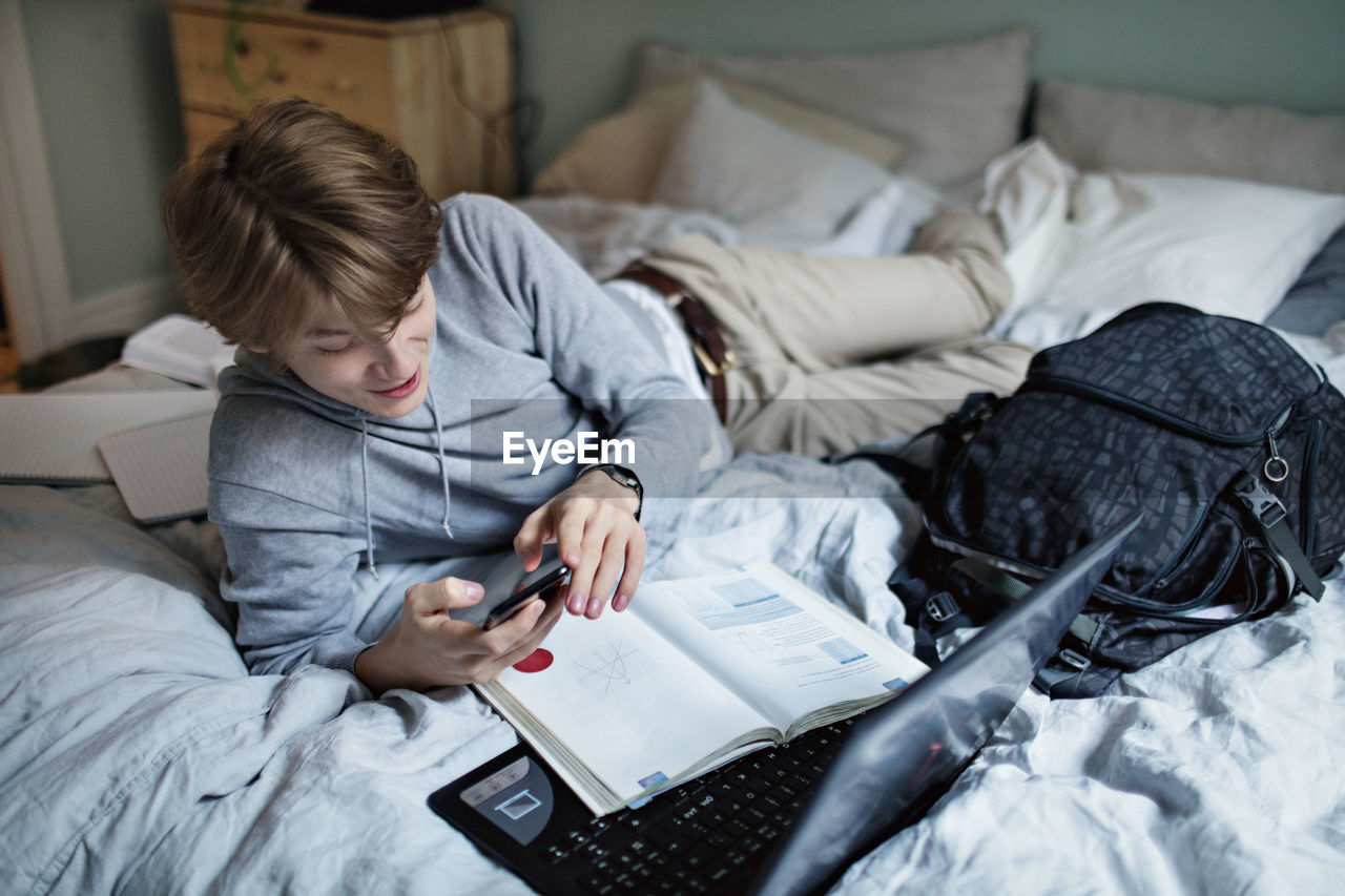 Addictive teenage boy using social media on mobile phone while doing homework in bedroom