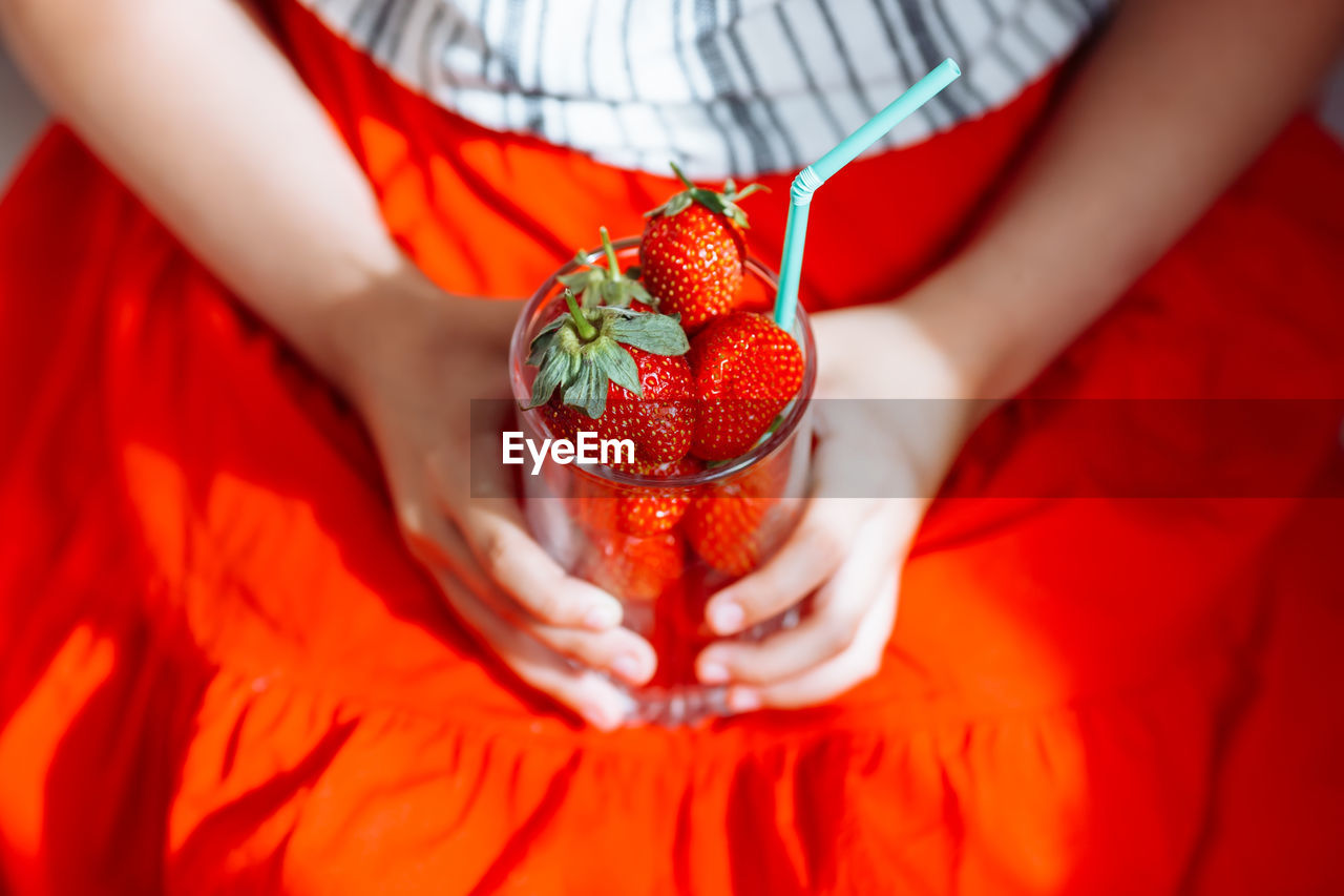 The hands of a girl in a red skirt hold a glass with juicy strawberries.