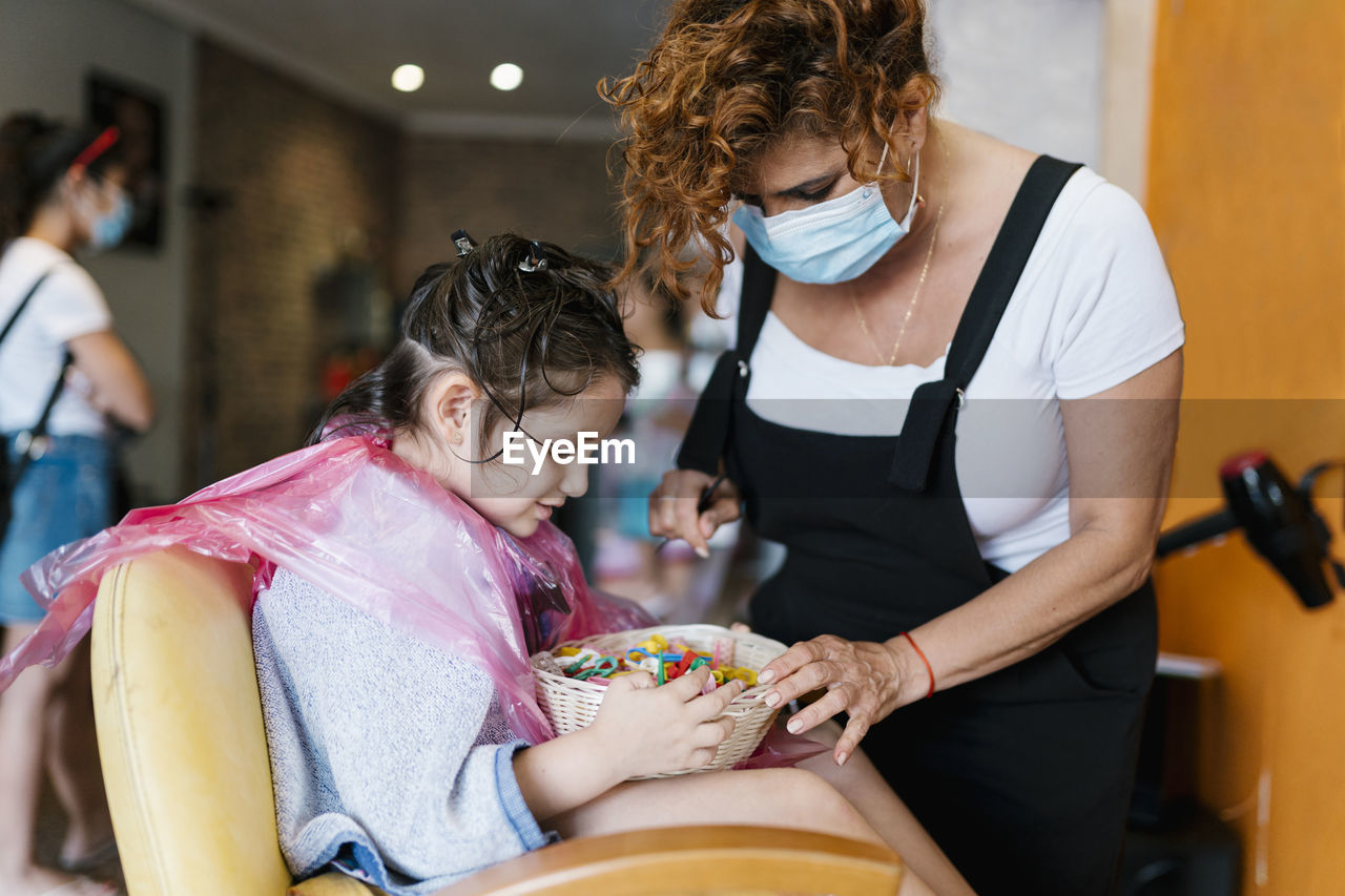 Hairdresser and girl searching in basket with hair clips at barber shop during coronavirus
