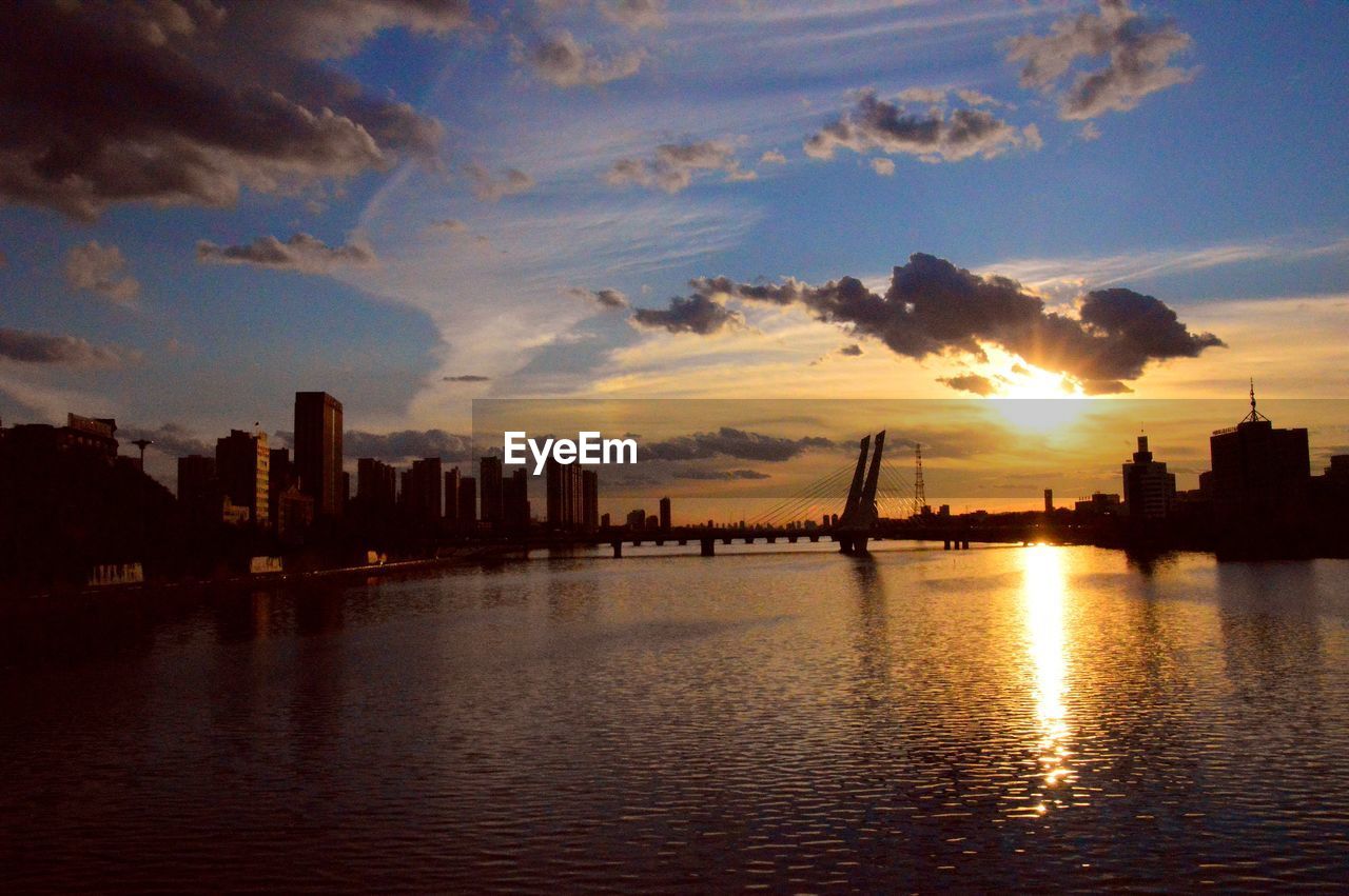 SILHOUETTE BUILDINGS BY RIVER AGAINST SKY DURING SUNSET IN CITY