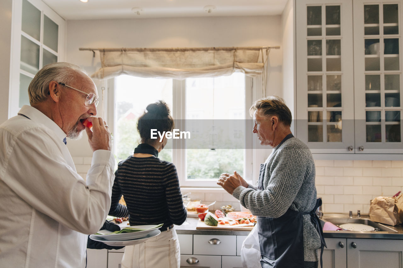 Man eating watermelon while friends talking in kitchen