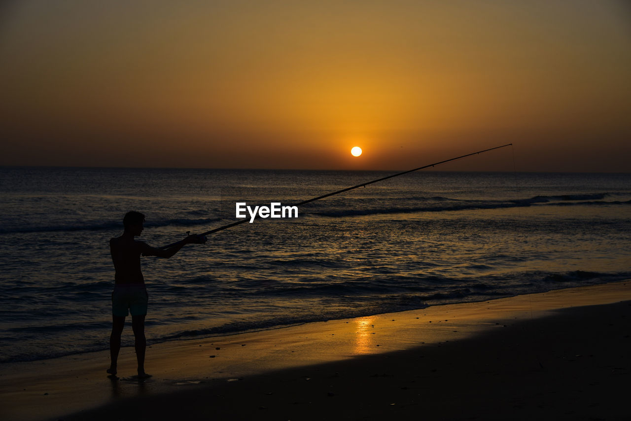 Silhouette man fishing at beach during sunset