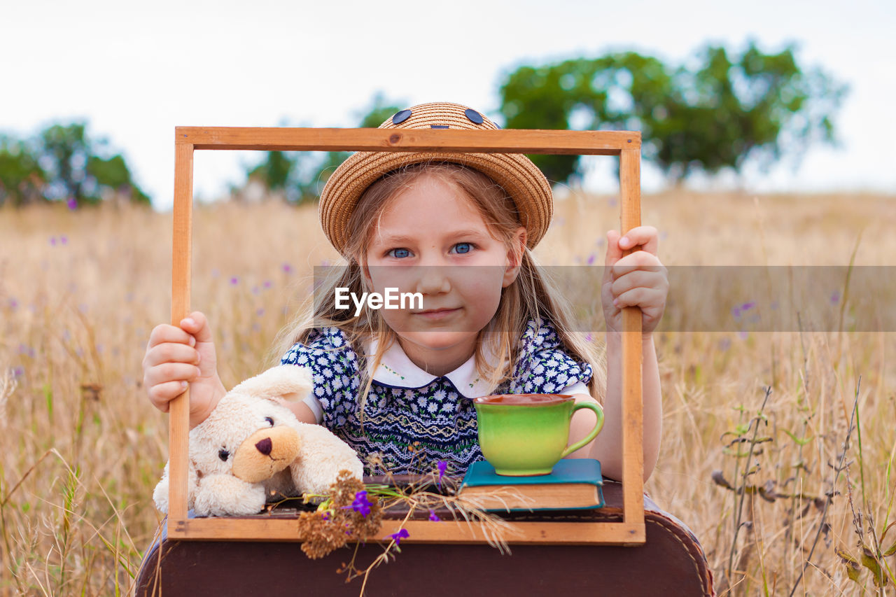 Child girl in straw hat vintage suitcase with cup tea cute kid with soft toy looking in wooden frame