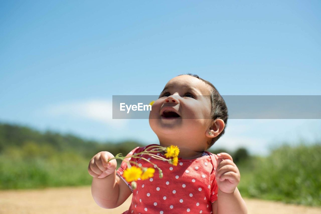 Close-up of baby girl holding yellow flowers against blue sky