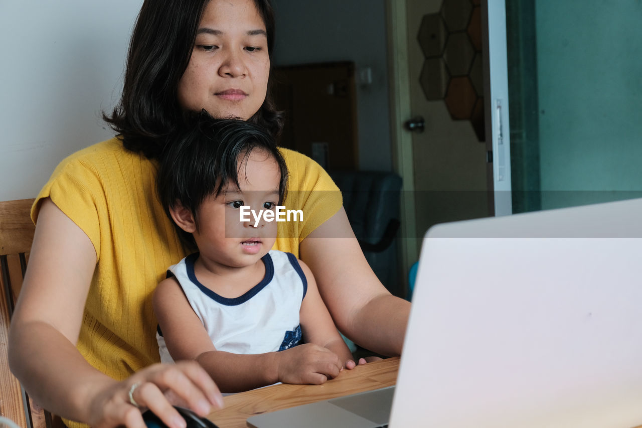 REAR VIEW OF MOTHER AND DAUGHTER SITTING ON LAPTOP AT HOME