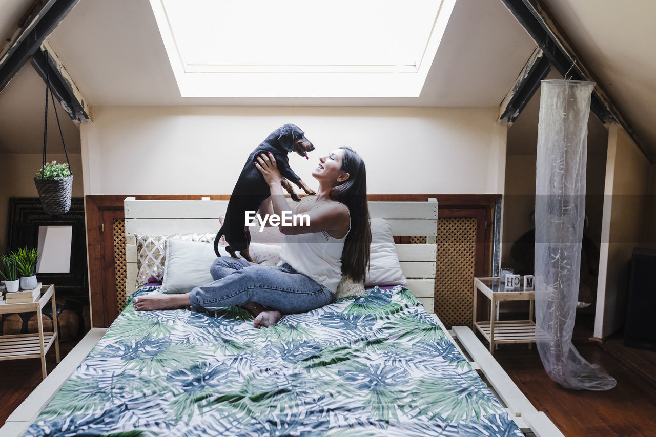 Playful woman holding dog while sitting on bed at home