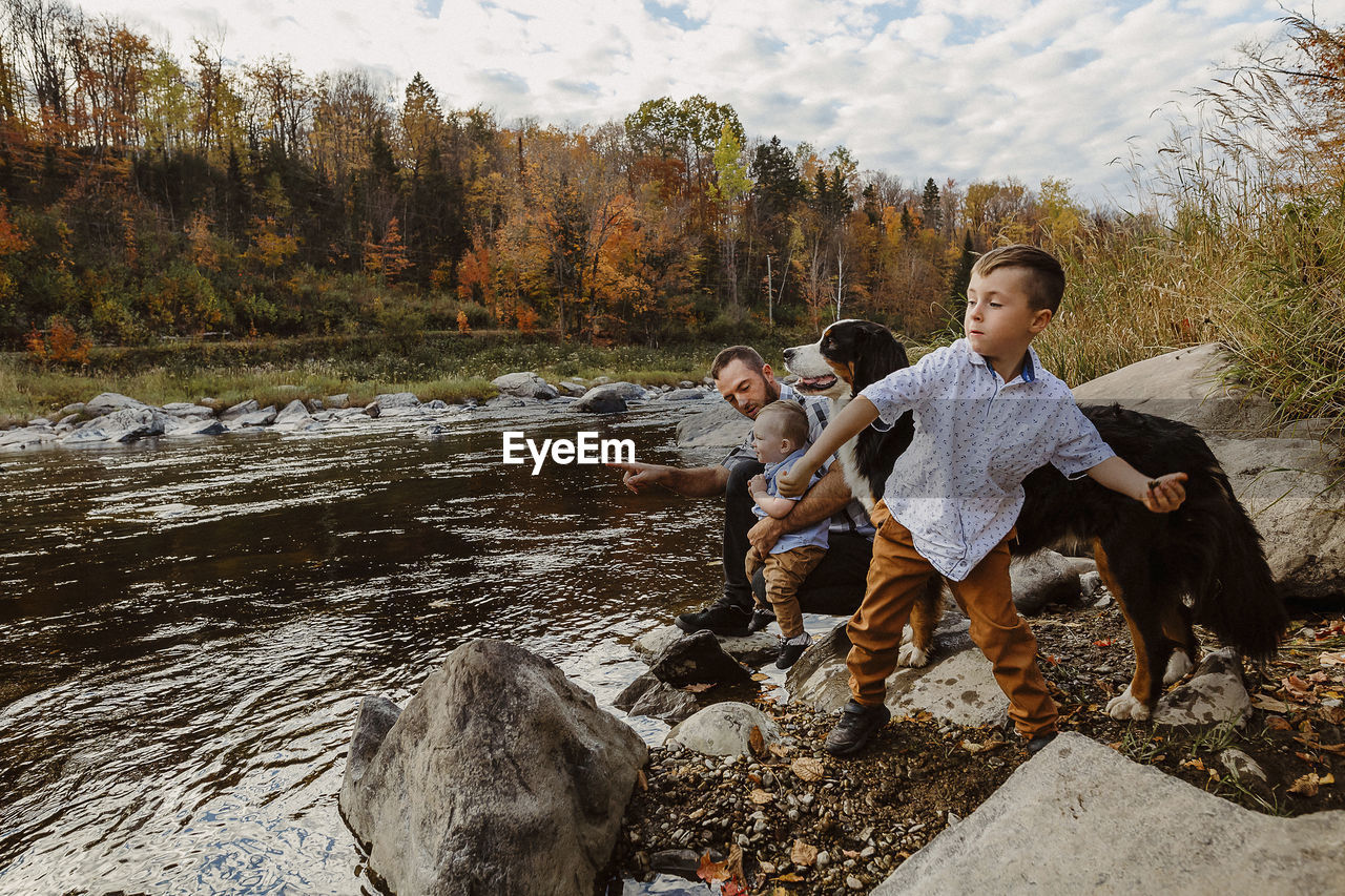 Father with sons and dog enjoying by river against cloudy sky in forest