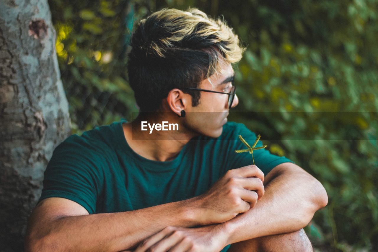 Young man looking away while sitting by tree trunk