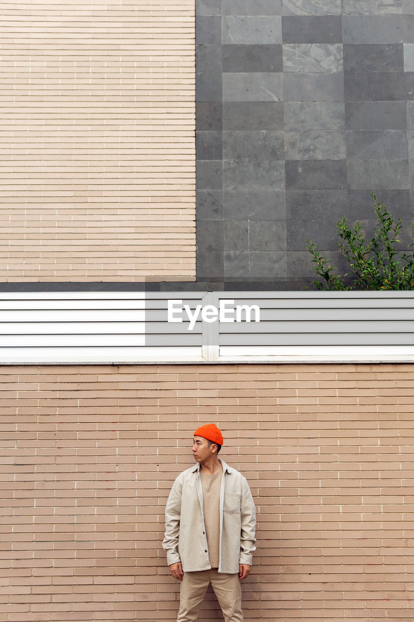 Calm serious stylish male in casual outfit standing against masonry wall looking way