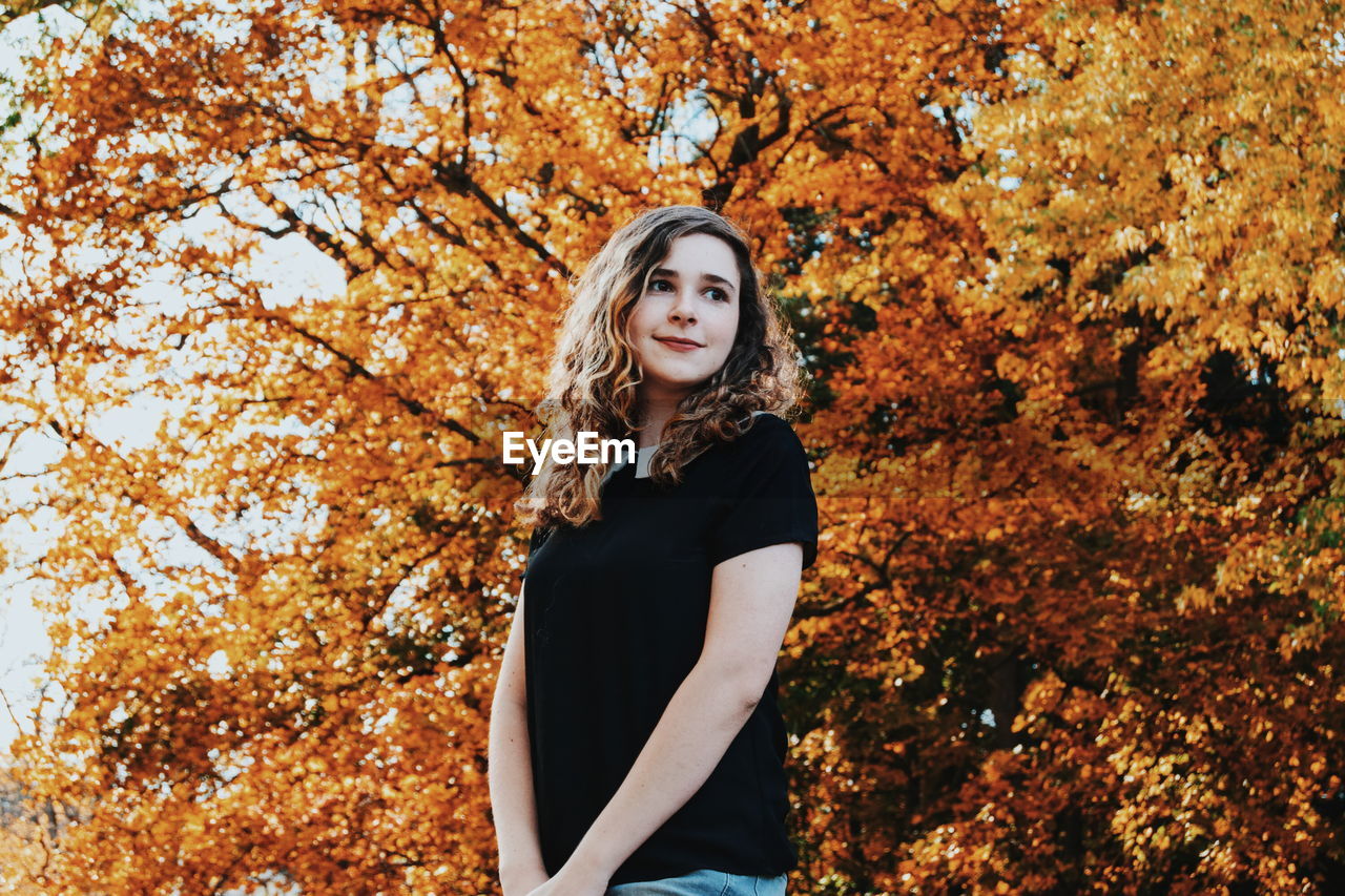 Low angle view of smiling woman standing against trees during autumn