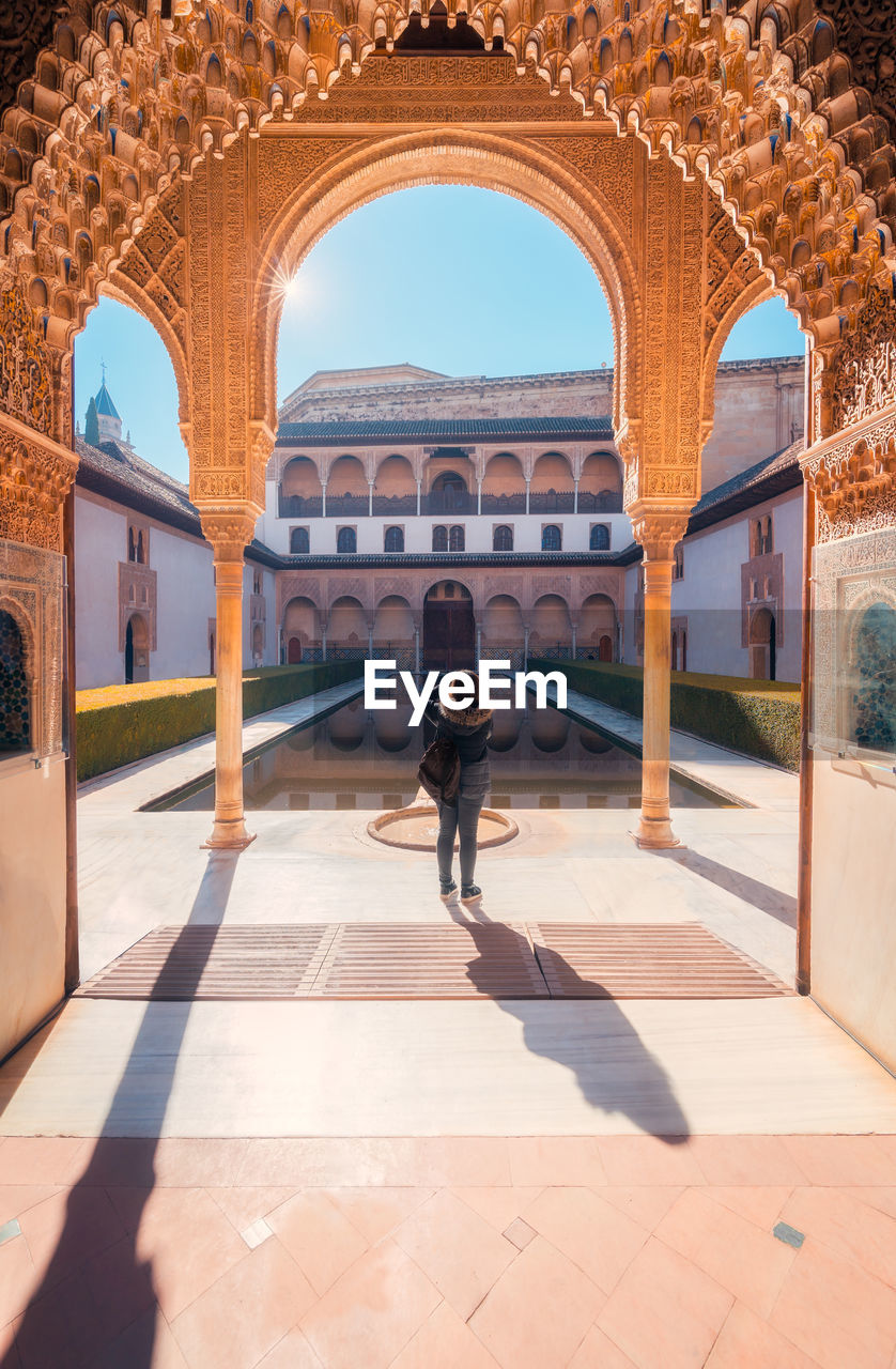 Anonymous woman standing under arch near calm pool and shooting sunlit court of the myrtles of comares palace on weekend day in granada, spain