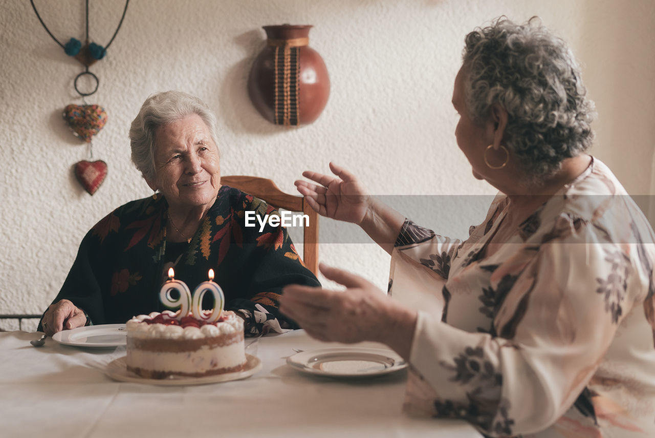 Elderly woman with gray hair and senior female sitting at dining table and celebrating 90th birthday with delicious cake with candles