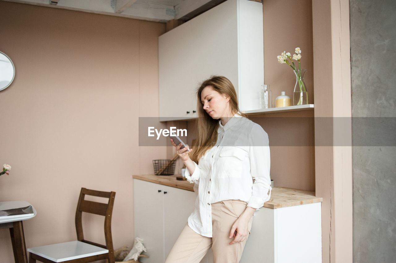 Young woman in white shirt looks to smart phone in home kitchen.