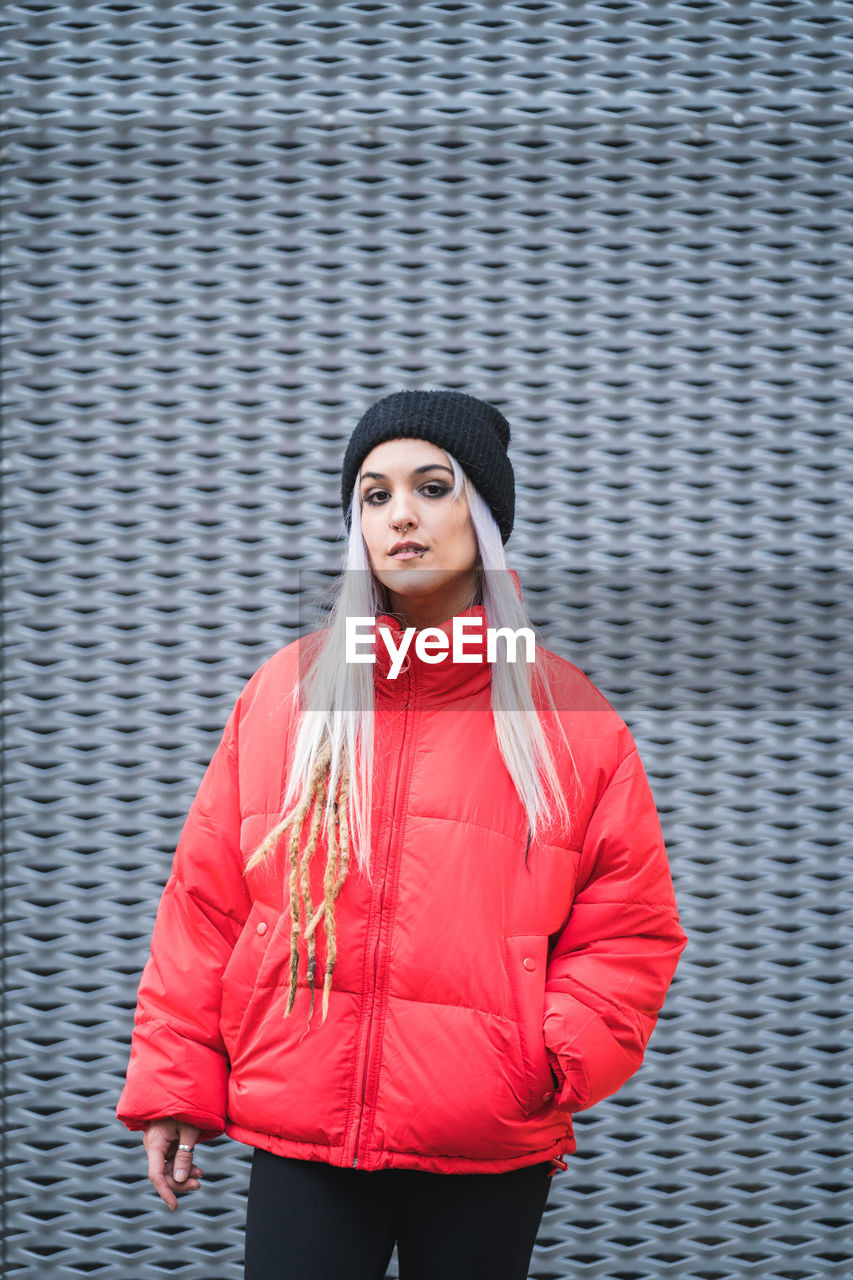 Blonde young hipster woman in stylish red warm jacket and hat standing against modern metal wall background