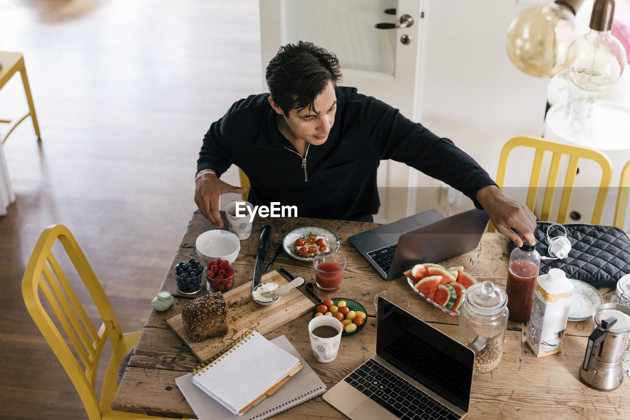 High angle view of male freelancer having breakfast while sitting with laptop on dining table at home