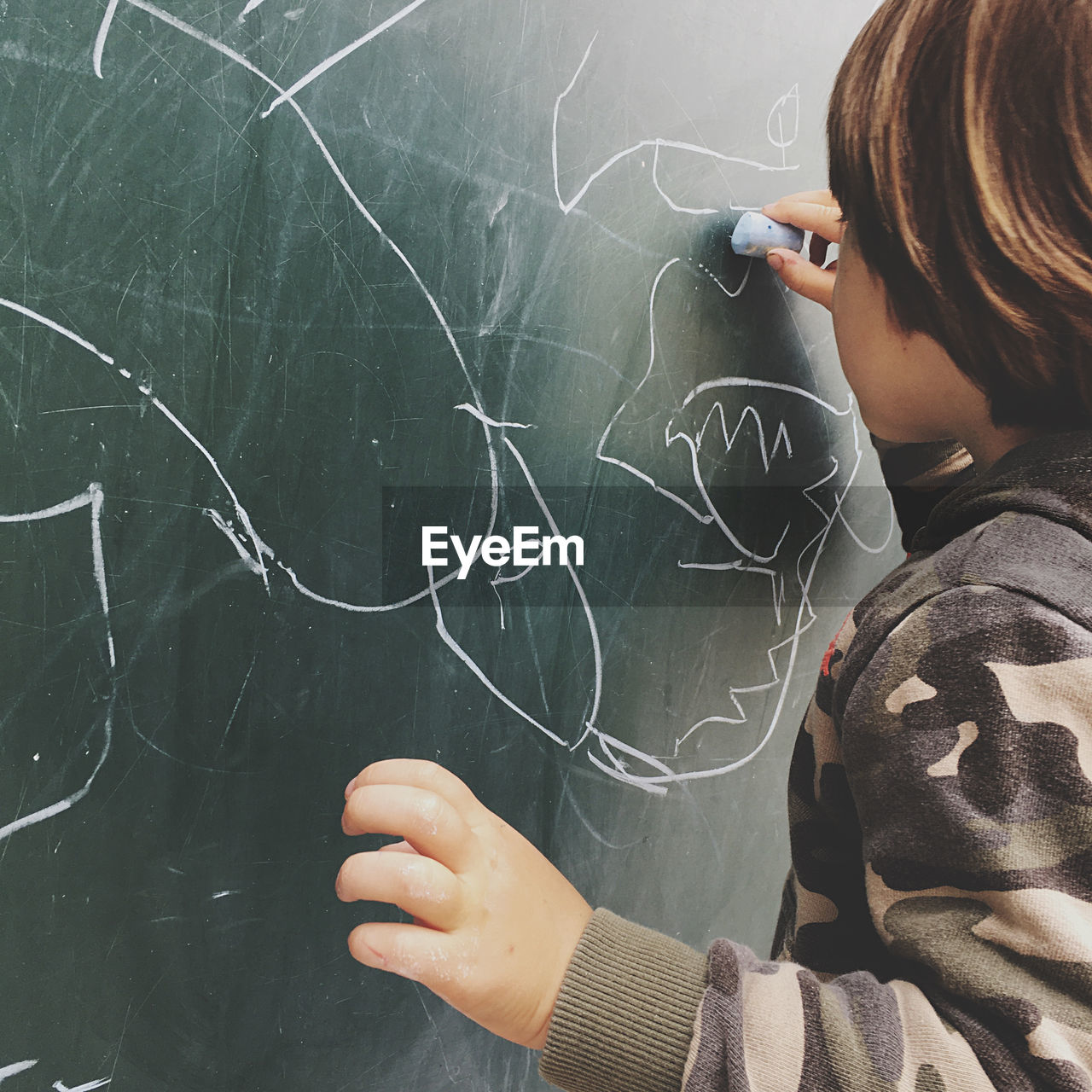 Rear view of boy drawing on blackboard at classroom