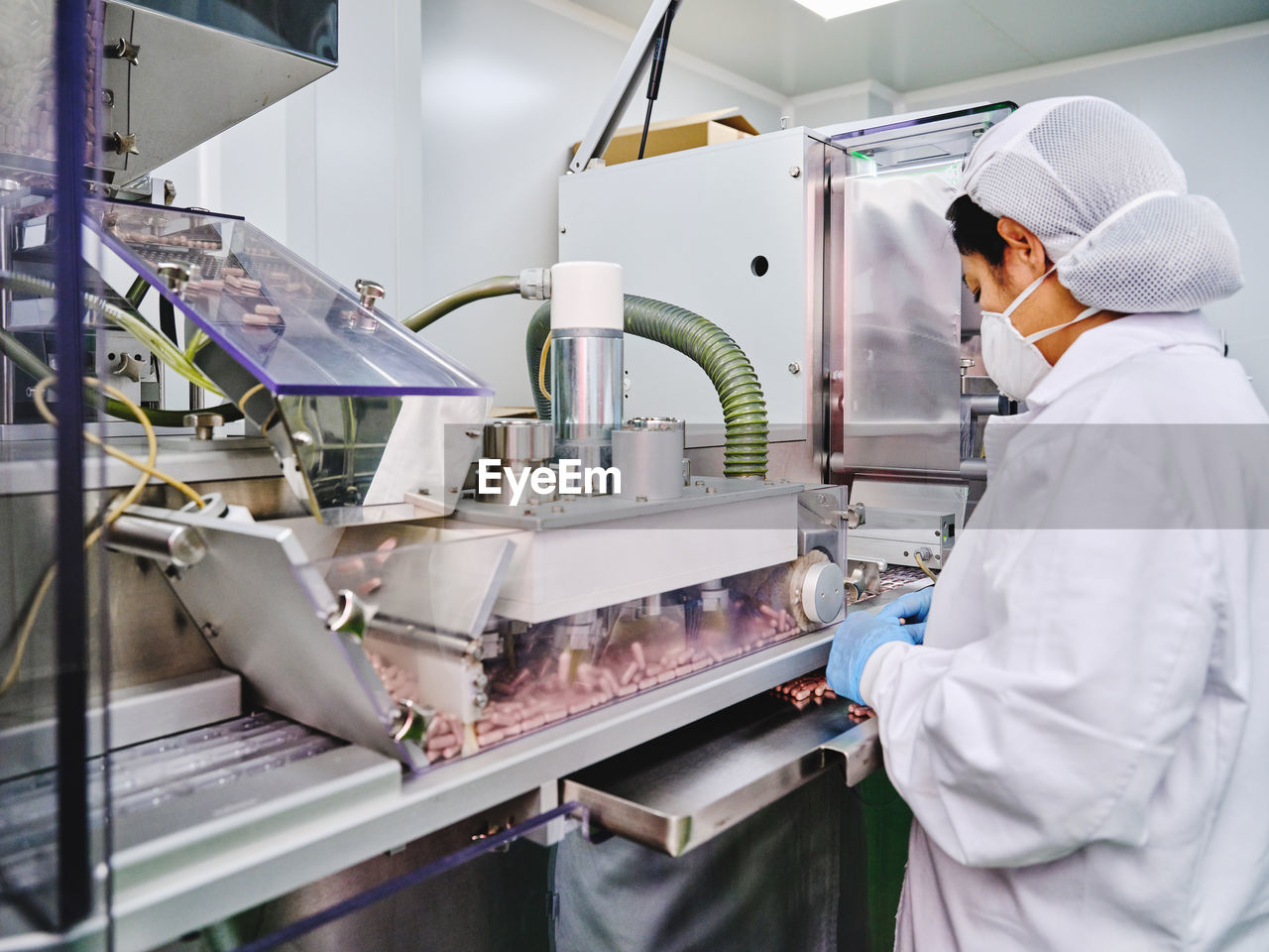 Side view of female factory worker in uniform operating conveyor with pills in pharmaceutical manufacturing laboratory