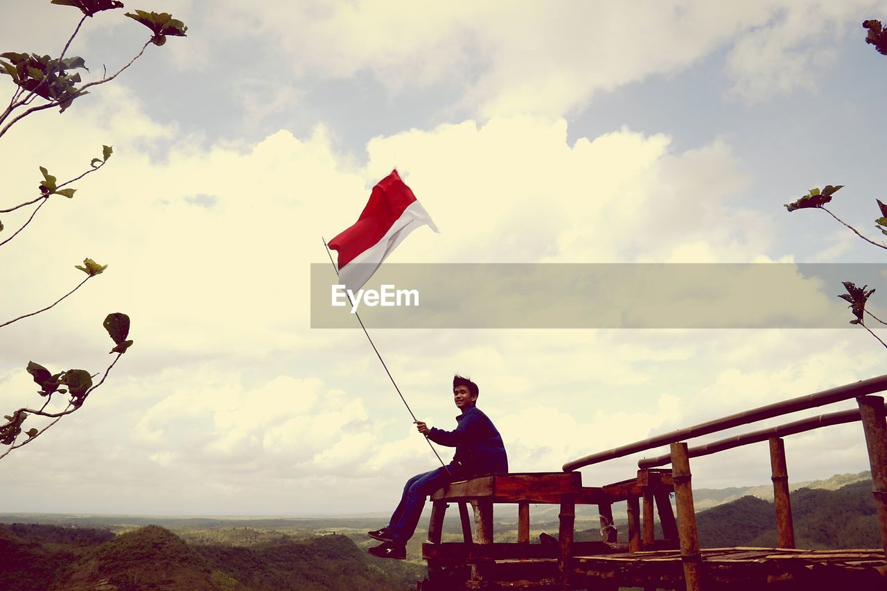 Smiling man with indonesia flag sitting on observation point over mountain against sky