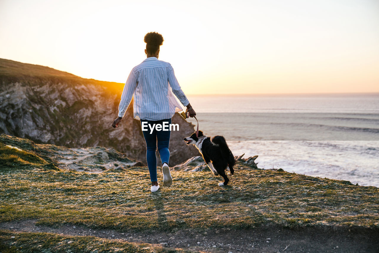 Back view of unrecognizable african american female owner running with border collie dog while spending time together on beach near waving sea at sunset