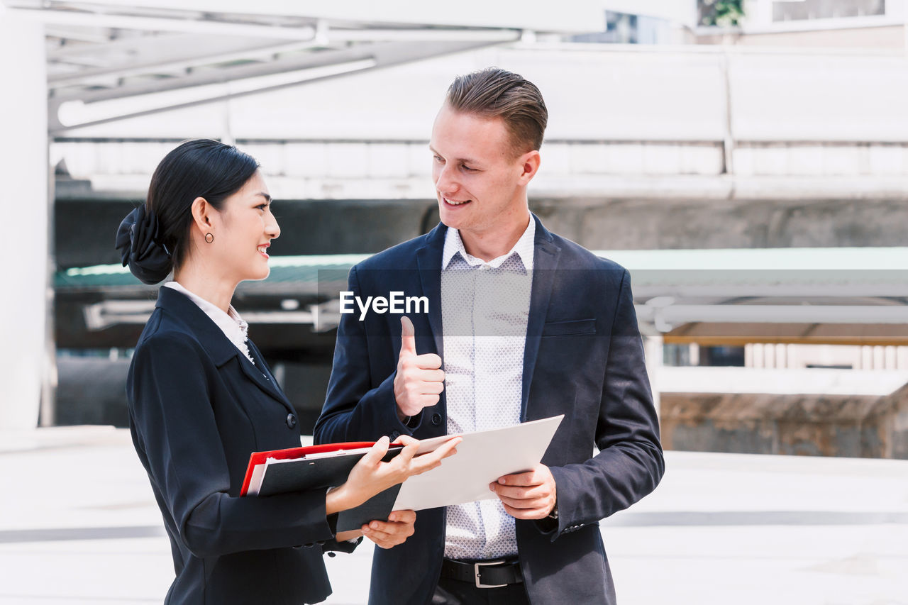 Smiling businessman showing thumbs up to colleague standing with data against building 