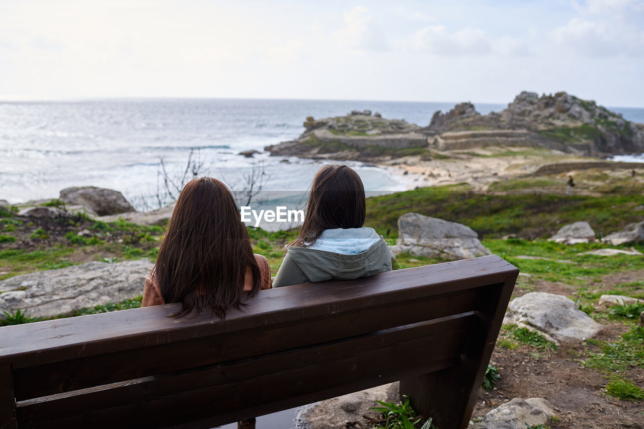 Rear view of women sitting on bench by sea