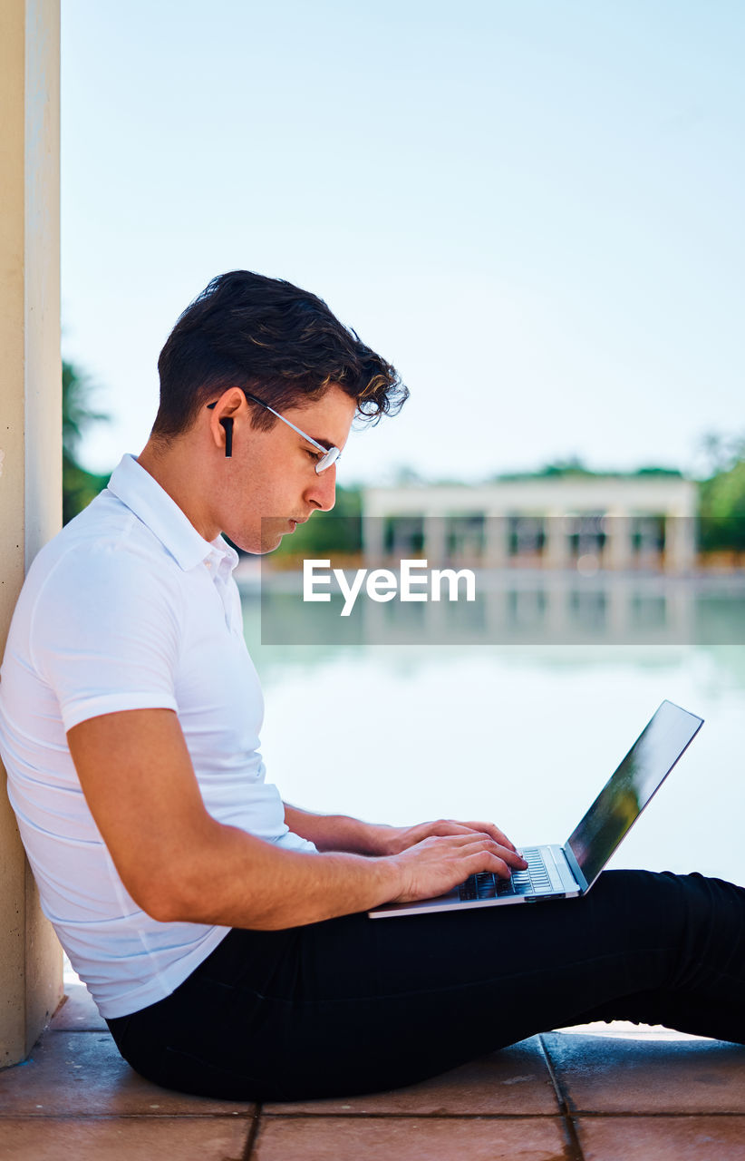 Side view of concentrated male student browsing netbook while sitting near stone column in university campus