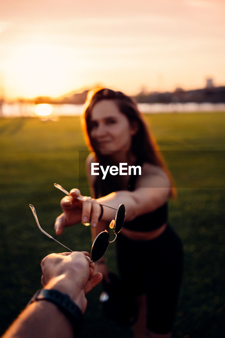 Young woman holding hands on field against sky during sunset