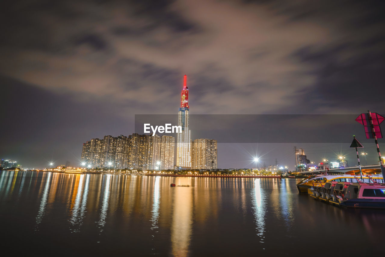 ILLUMINATED BUILDINGS BY RIVER AGAINST SKY