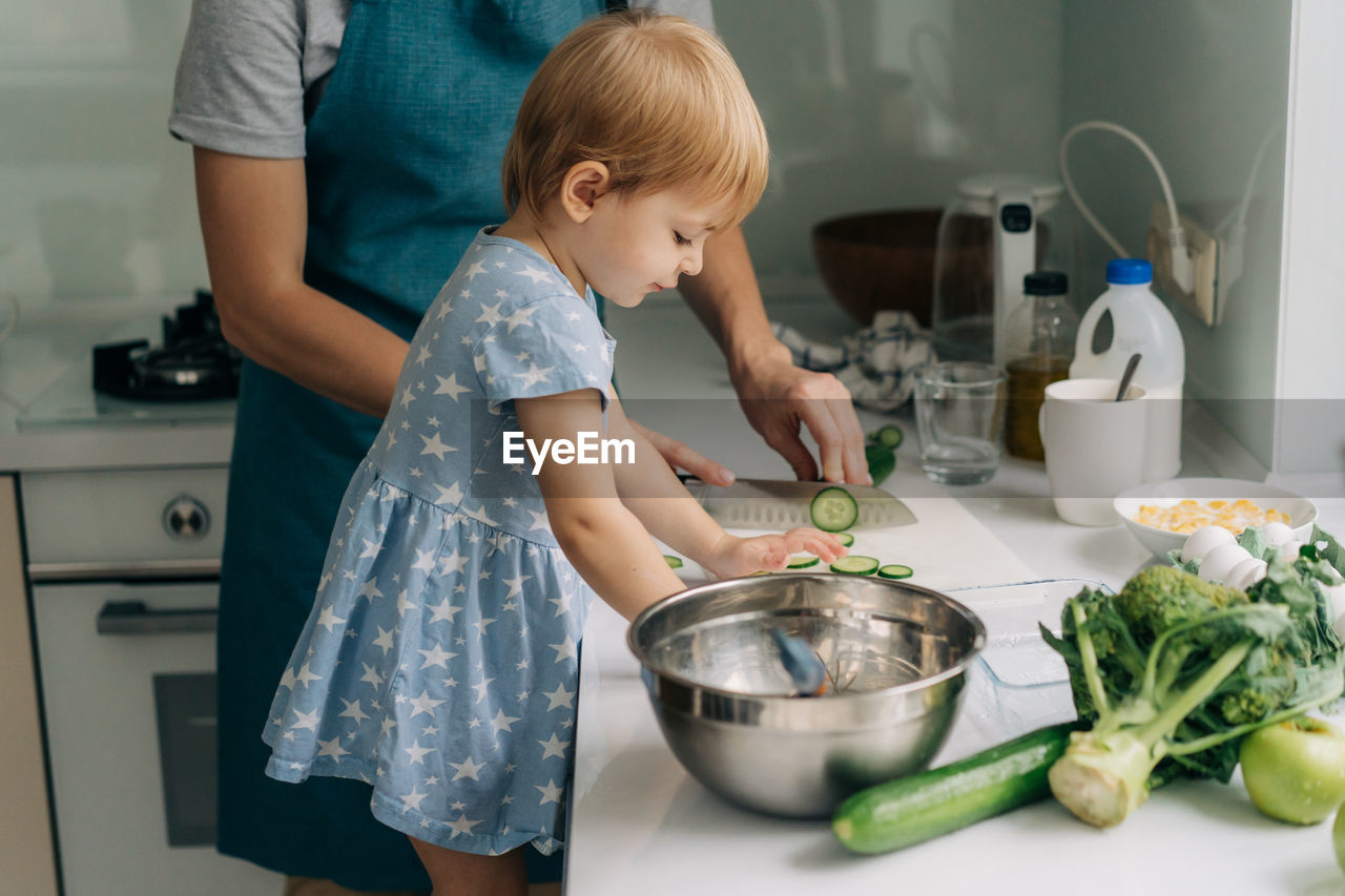 Toddler daughter helps mom cook in the home kitchen.