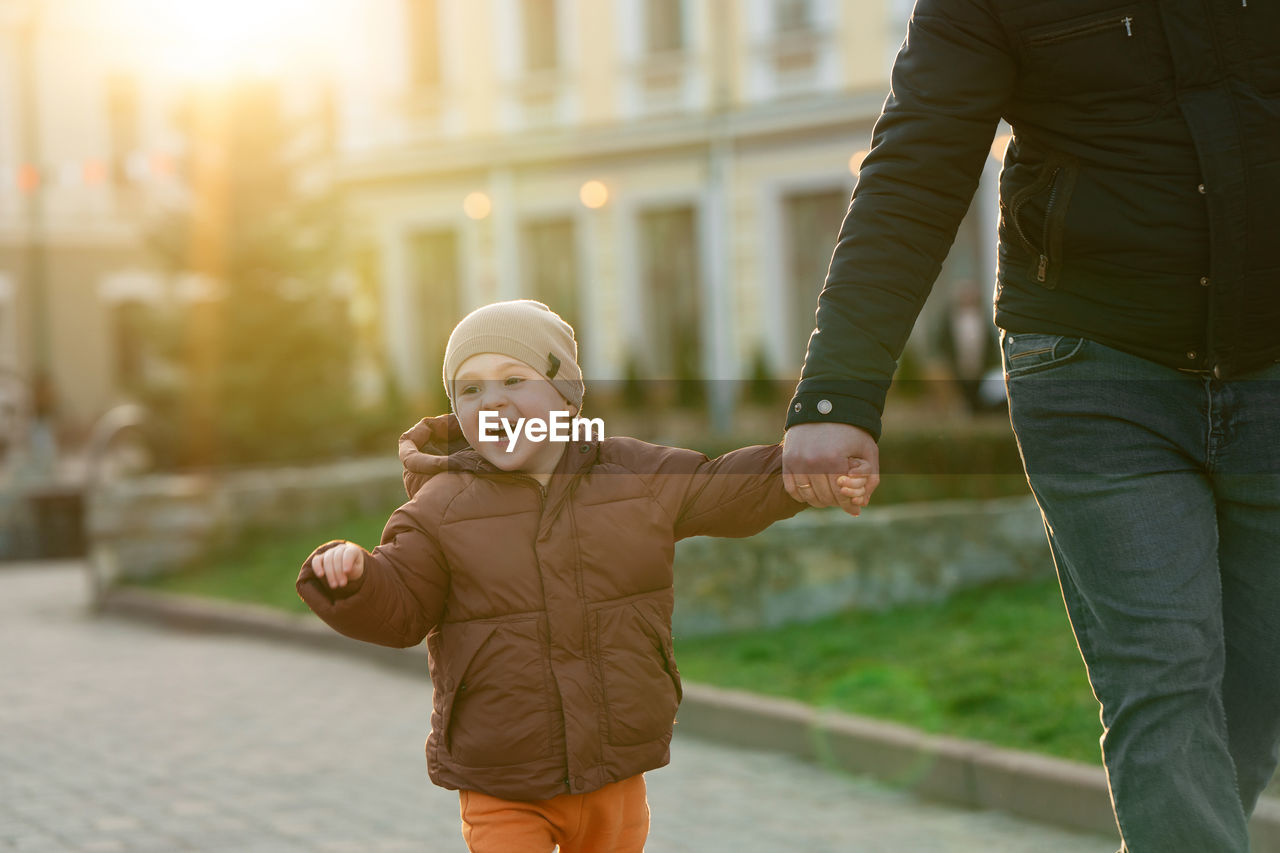 A happy three year old son walks hand in hand with his dad in the city on a sunny spring day. 