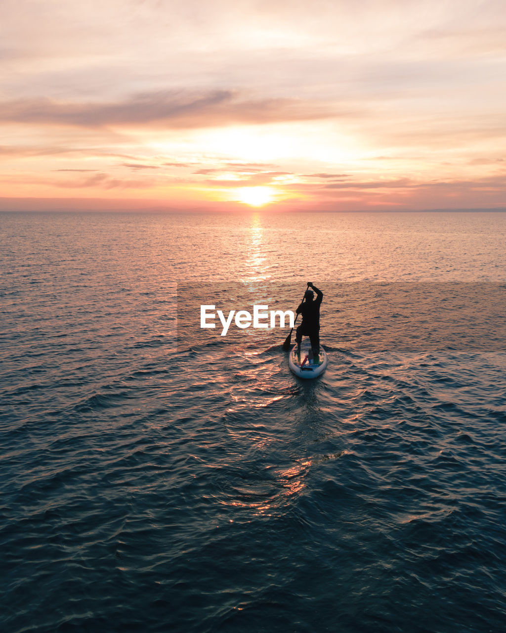 A man surfing  on a board with paddle into the sunset