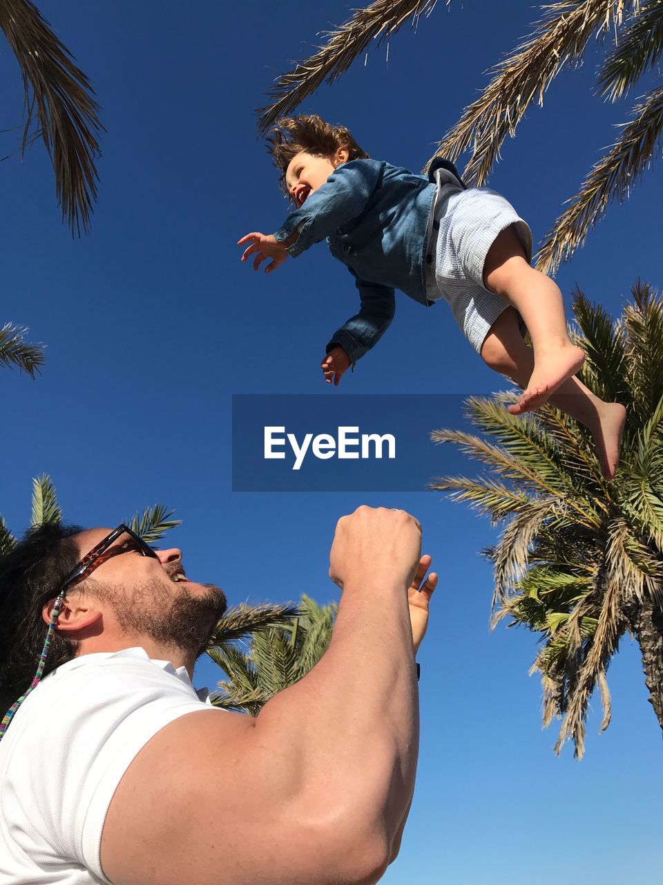 Low angle view of man playing while throwing boy against trees and clear blue sky