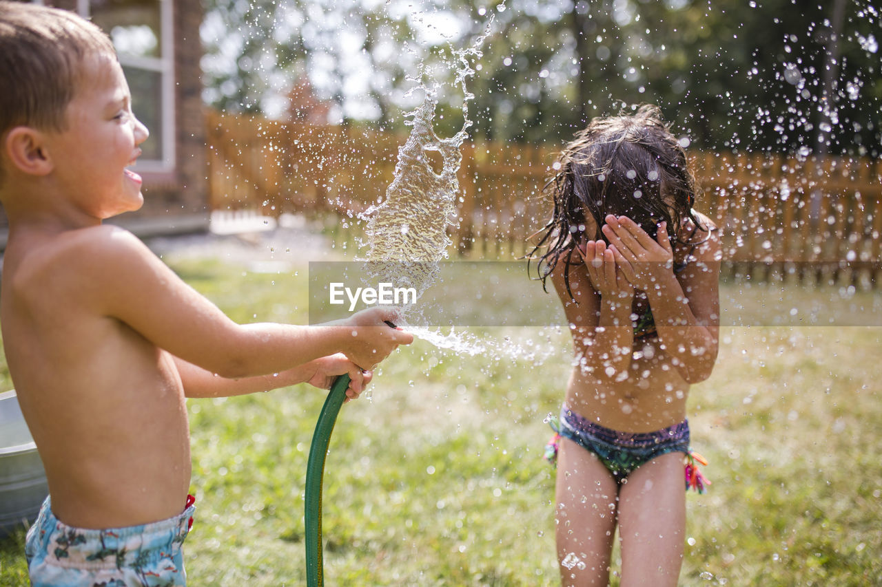 Shirtless brother spraying water on sister through garden hose at yard