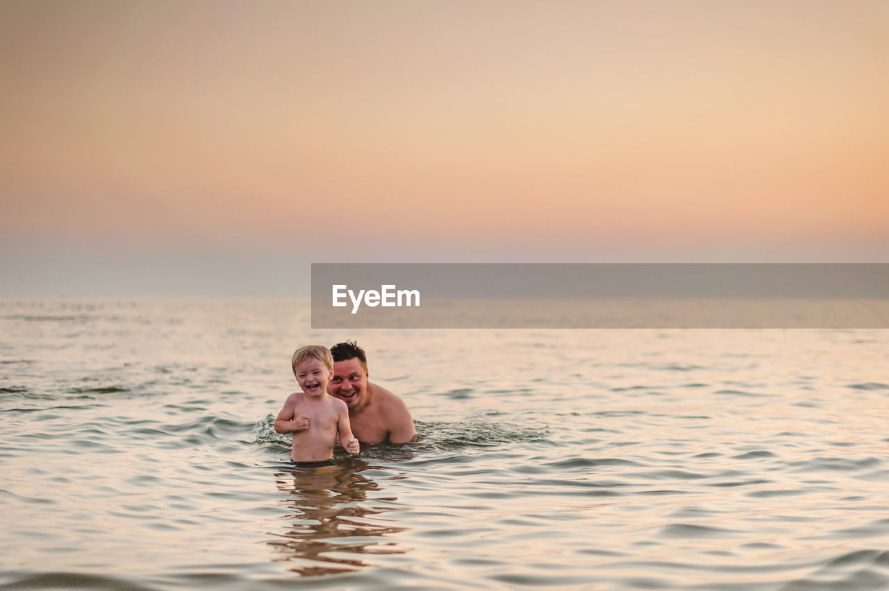 Portrait of couple swimming in sea against sky during sunset