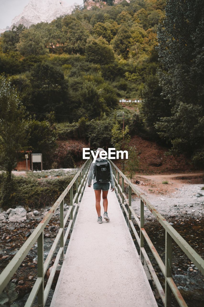 REAR VIEW OF WOMAN WALKING ON FOOTBRIDGE AMIDST TREES