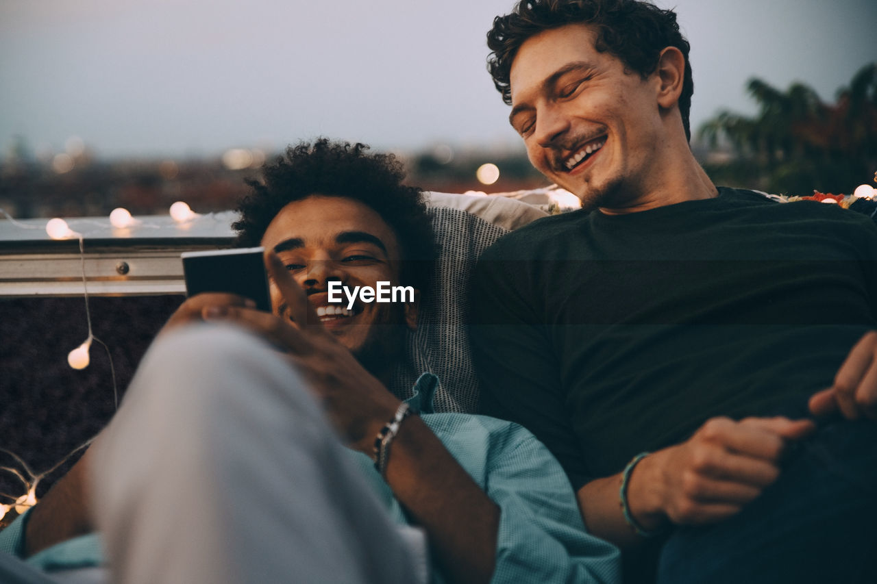 Happy male friends looking at smart phone relaxing on terrace during rooftop party in city