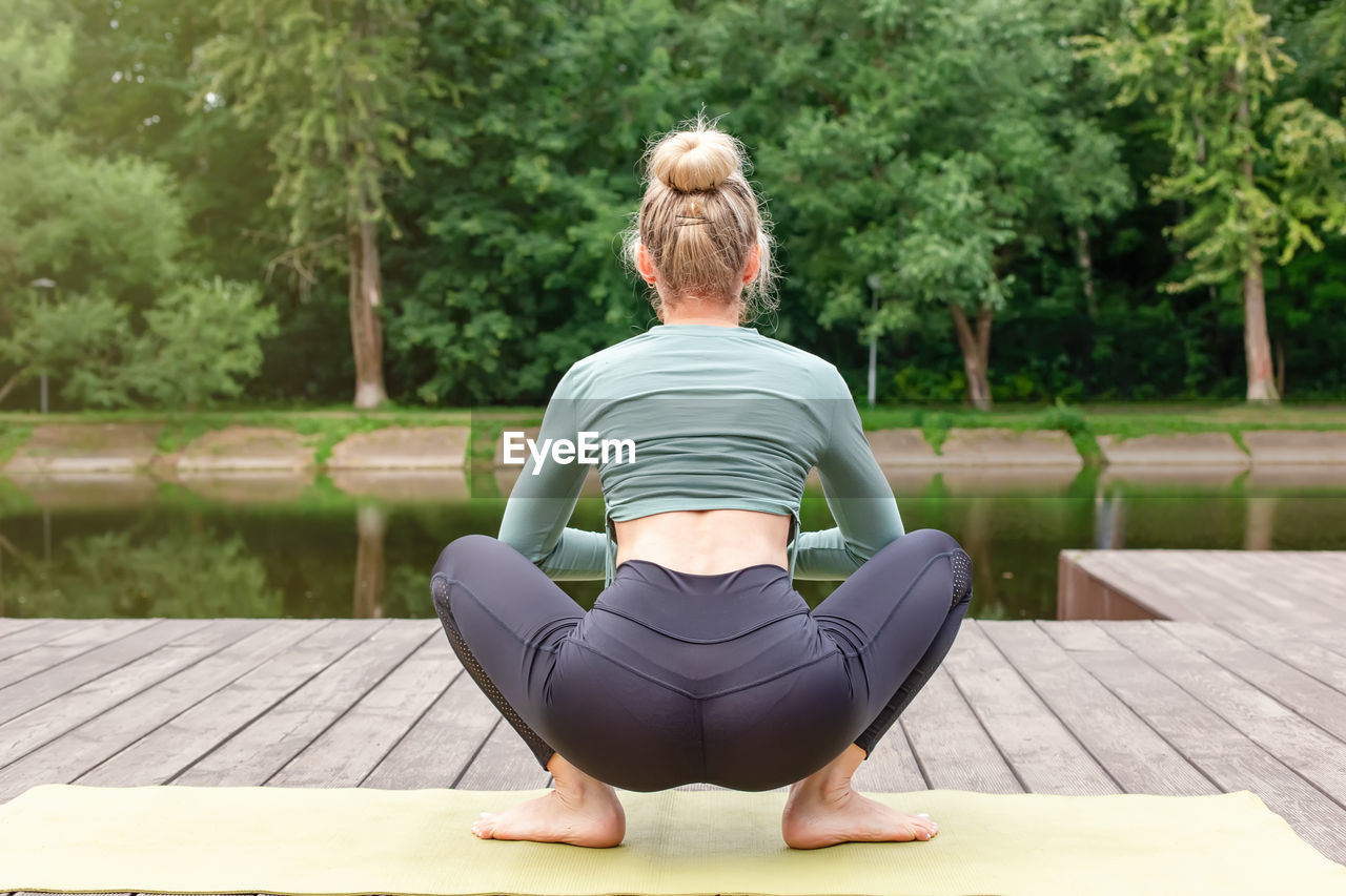 A slender woman sitting on a wooden platform by a pond on green mat in summer, does yoga