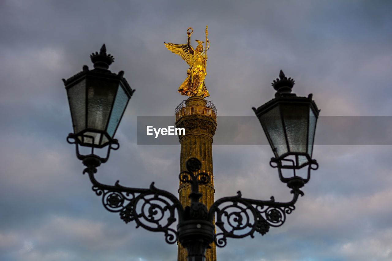 Low angle view of victory column and street light against cloudy sky